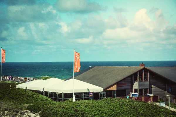 Ein Blick auf das Meer, der das Restaurant Strandoase mit einer großen Holzterrasse und weißen, mit Baldachinen überdachten Sitzbereichen zeigt. Zwei orangefarbene Flaggen mit Text wehen und hinter dem Restaurant erstreckt sich der weite blaue Ozean unter einem teilweise bewölkten Himmel bis zum Horizont. Der Sandbereich, der zum Strand führt, ist von Grün umgeben.