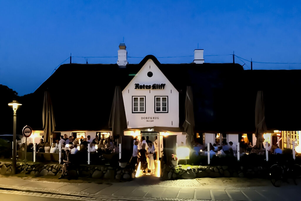 Ein gemütliches Restaurant namens „Rotes Kliff Dorfkrug“ mit Strohdach, das in der Abenddämmerung warm leuchtet. Die Gäste sitzen unter dunklen Sonnenschirmen und genießen ihr Essen. Der Himmel ist tiefblau und Kopfsteinpflasterwege mit Außenbeleuchtung umgeben den Sitzbereich des Dorfkrugs und schaffen eine einladende Atmosphäre.