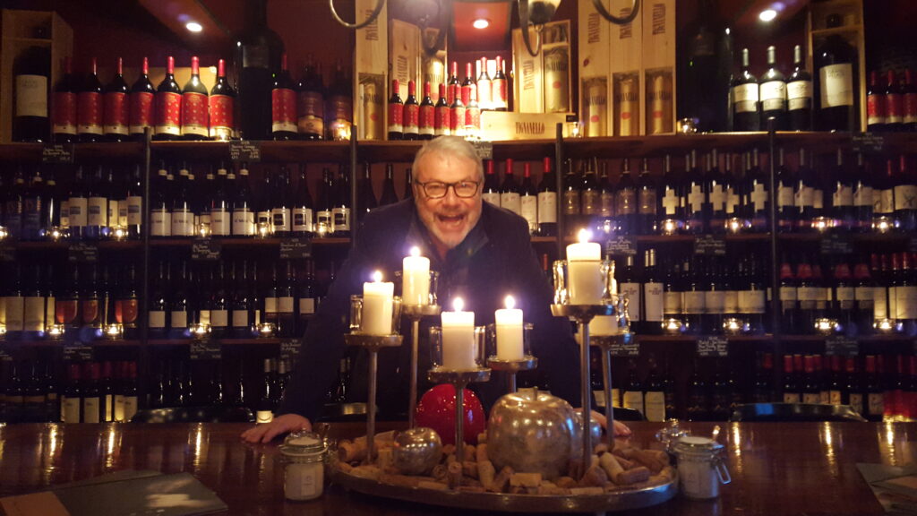 Ein Mann mit Brille, Pius, steht hinter einer Bar, die mit brennenden Kerzen und Dekoartikeln geschmückt ist. Regale voller Weinflaschen bilden den Hintergrund und schaffen eine gemütliche, intime Atmosphäre. Pius lächelt und scheint die Atmosphäre der Umgebung zu genießen.