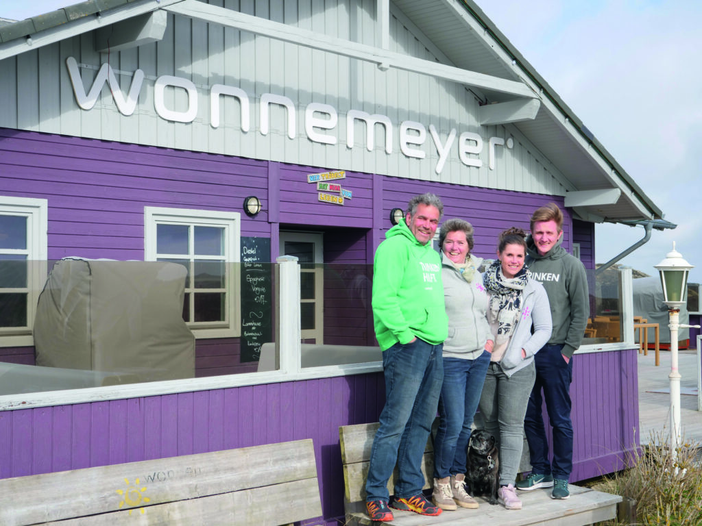 Vier Menschen, alle leger gekleidet, stehen vor einem lilafarbenen Gebäude, auf dem in großen weißen Buchstaben „Wonnemeyer“ steht, und lächeln herzlich. Zwei Männer und zwei Frauen stehen auf einer Holzterrasse und haben einen Koffer vor sich. Das Gebäude hat ein schräges Dach, hellgraue Zierleisten und eine Außenlaterne.