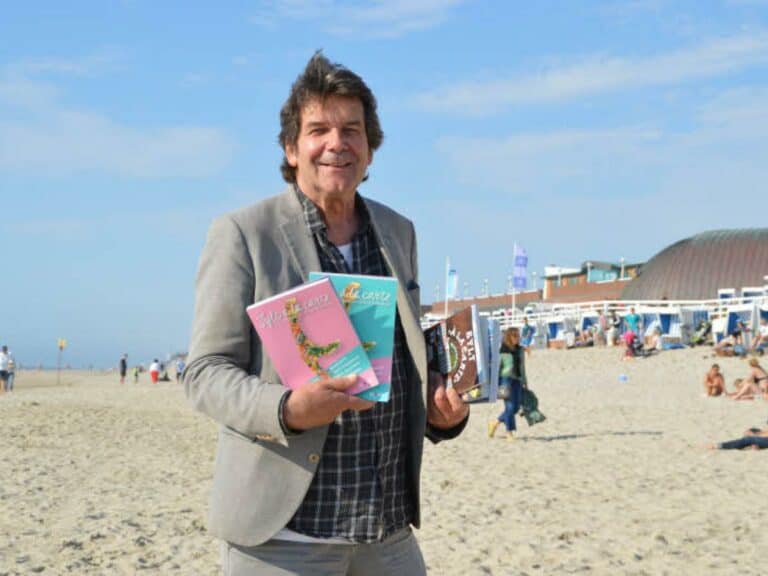 Ein Mann in einem grauen Blazer und einem schwarzen Hemd steht lächelnd an einem Sandstrand und hält drei bunte Bücher hoch. Der Strand ist von Menschen gesäumt, die den sonnigen Tag genießen. Im Hintergrund ist ein Gebäude mit blauen Flaggen zu sehen, über ihm ist ein klarer Himmel.