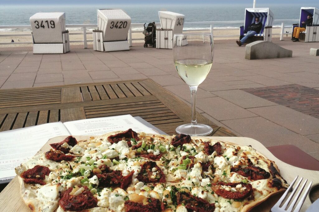 Ein Tisch mit Blick auf einen Strand mit weißen Strandhütten im Hintergrund. Auf dem Tisch liegt ein offenes Buch, eine dünne Pizza mit sonnengetrockneten Tomaten, Feta und Kräutern und ein Glas Weißwein. In der Ferne entspannen Freunde in Strandstühlen mit Blick aufs Meer.