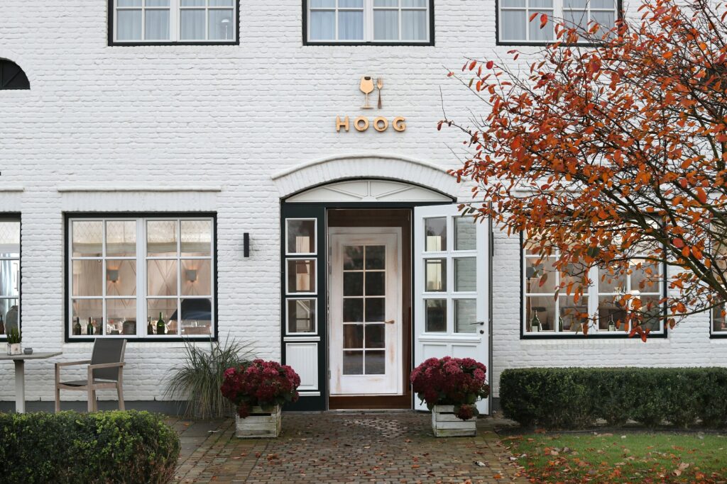 Ein weißes Backsteinhaus mit einer Glastür und zwei großen Fenstern auf beiden Seiten. Über der Tür steht auf einem Holzschild „Hoog“. Rote Blumentöpfe schmücken beide Seiten des Eingangs, und auf der linken Seite steht ein kleiner Tisch mit Stühlen. Auf der rechten Seite des Weges steht ein Baum mit orangefarbenen Blättern.