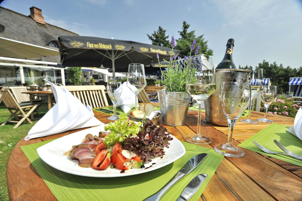 Ein für ein Essen im Freien gedeckter Holztisch ist mit einem Teller Salat und dünn geschnittenem Fleisch geschmückt, umgeben von Weingläsern, einem Topf mit Kräutern, Besteck auf grünen Servietten und einer Champagnerflasche in einem Eiskübel. Im Hintergrund genießen Lee und Hus die von schwarzen Sonnenschirmen und Grünpflanzen beschattete Restaurantterrasse.