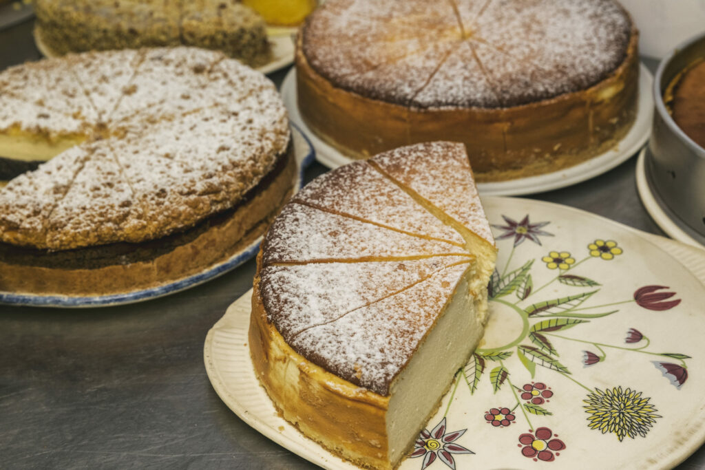 Auf einem Tisch in der Kleinen Teestube werden verschiedene Käsekuchen präsentiert. Zwei sind auffällig mit Puderzucker bestreut und liegen teilweise angeschnitten auf dekorativen Tellern. Einer hat einen Teller mit detailliertem Blumenmuster, während die anderen Teller schlichter gehalten sind. Im Hintergrund sind weitere ganze und angeschnittene Kuchen zu sehen.