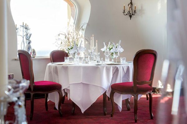 Ein runder Tisch ist elegant für eine Mahlzeit mit einer weißen Tischdecke, Kristallgläsern und weißen Blumendekorationen gedeckt. Vier rot gepolsterte Holzstühle umgeben den Tisch. Ein Kandelaber und ein Kronleuchter werten das klassische Dekor im Miramar-Raum auf, der über ein Fenster und weiße Wände verfügt.