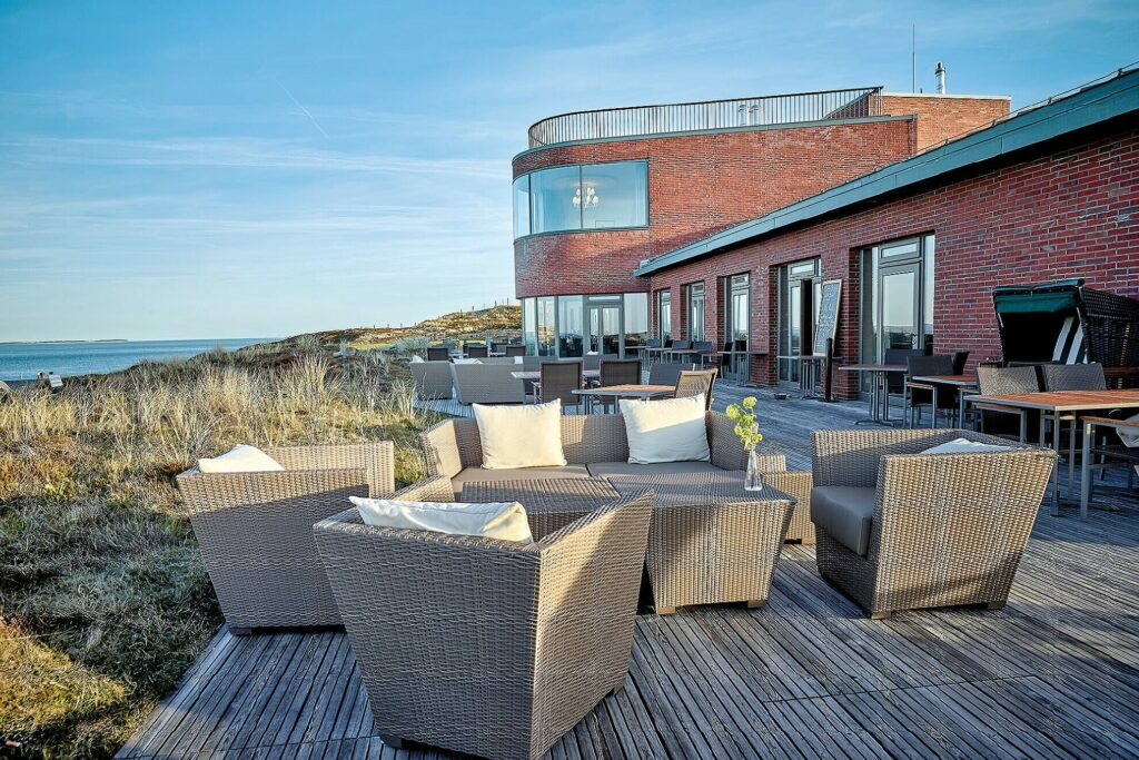 Außenterrasse mit Korbmöbeln von Strönholt, darunter Sofas und Sessel, auf einem Holzdeck mit Blick auf eine malerische Küstenlandschaft. An einer Seite grenzt ein modernes, geschwungenes Backsteingebäude mit großen Fenstern. Der Himmel ist klar und blau und verstärkt die ruhige Atmosphäre. In der Nähe des Gebäudes stehen Tische mit Stühlen.