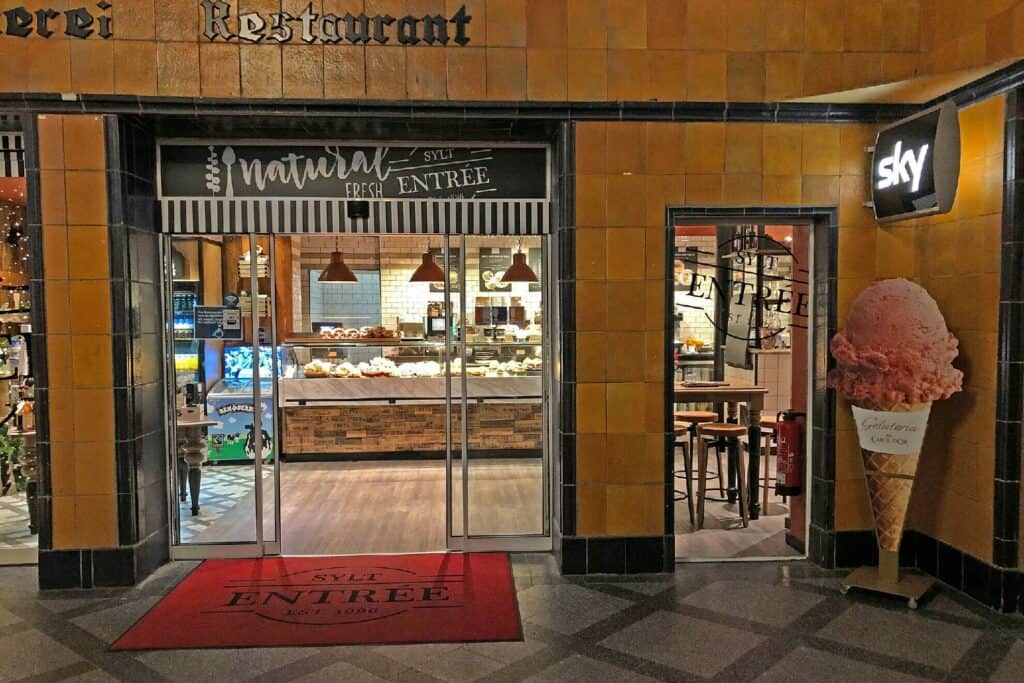 Eingang zu einer hell erleuchteten Bäckerei und einem Restaurant. Auf dem Schild darüber steht „Natural Fresh Entree“. Im Inneren befindet sich eine Theke mit Lebensmitteln und Hängelampen. Rechts markiert ein Eisdekor mit einer großen Tüte den Eingang zum Sylter Eisbereich mit einem „Himmel“-Schild darüber.