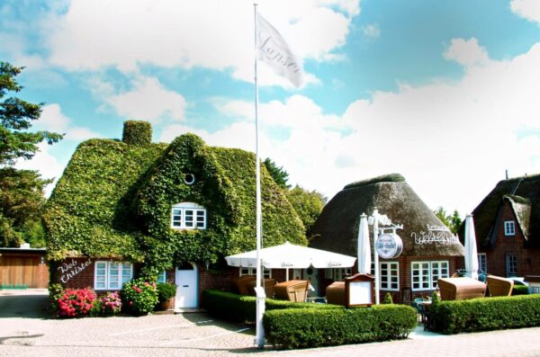 Eine malerische Szene mit zwei charmanten, efeubewachsenen Cottages mit Strohdächern, die an eine Illustration von Web Christel erinnern. Zwischen den Gebäuden weht eine weiße Flagge. Sitzgelegenheiten im Freien mit Sonnenschirmen lassen an ein Café oder Restaurant denken. Die Umgebung ist sonnig mit einem klaren blauen Himmel und leichten Wolken.