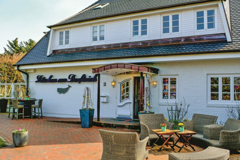 Ein charmantes zweistöckiges weißes Haus, bekannt als Fitschen am Dorfteich, verfügt über eine gemauerte Terrasse mit Korbstühlen, Topfpflanzen und Tischen im Freien. Das Gebäude hat ein Schild mit stilisiertem Text in der Nähe des Eingangs und ein dekoratives Vordach über einer modernen Tür. Fenster mit Fensterläden schmücken sowohl das Erdgeschoss als auch die oberen Stockwerke.