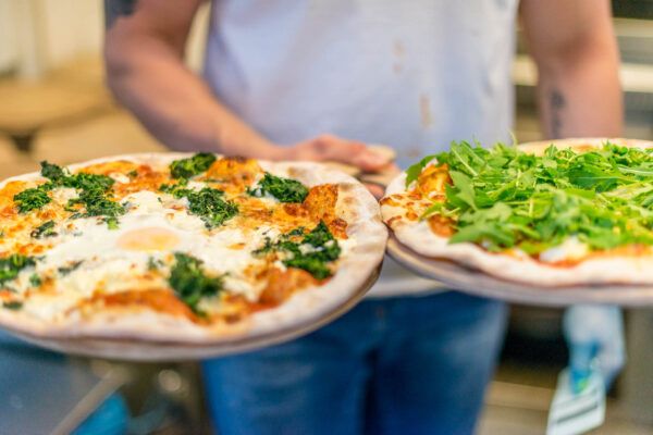 Eine Person in einem weißen Hemd hält zwei Pizzen, eine mit Gemüse und Käse belegt, die andere mit Rucola und Käse von Riva. Die Pizzen liegen auf Metallblechen, der Hintergrund ist leicht verschwommen, sodass die köstlichen Pizzen im Mittelpunkt stehen.