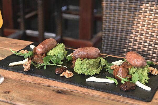 Ein Gourmetgericht, präsentiert auf einer dunklen Schieferplatte, bestehend aus drei Würstchen auf einem Haufen leuchtend grüner Erbsenpüree. Garniert wird die kulinarische Kreation mit Rucola, Walnüssen, Scheiben Weißkäse und einigen Pflaumen, alles kunstvoll und perfekt arrangiert im S-Point. Im Hintergrund sind ein Holztisch und Korbstühle zu sehen.