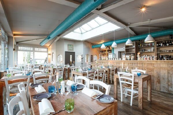 Ein gemütliches Restaurant-Interieur mit rustikalem Holzthema im Stadtgeflüster-Stil. Weiße Stühle und Holztische sind ordentlich angeordnet, akzentuiert durch blaue Gläser und Servietten. Der Barbereich verfügt über Regale voller Flaschen und ein Oberlicht sorgt für natürliches Licht. An der Decke sind freiliegende blaue Lüftungsschächte zu sehen.