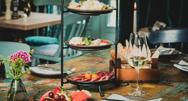 Auf einem rustikalen Holztisch steht ein dreistöckiger Ständer von Dorfkrug, auf dem verschiedene Käsesorten, Trauben und Brot liegen. Um den Ständer herum stehen Teller mit Obst und Fleisch, ein Weinglas mit Weißwein und eine Vase mit roten und weißen Blumen. Stoffservietten sind ordentlich platziert, während eine brennende Kerze für eine warme Atmosphäre sorgt.