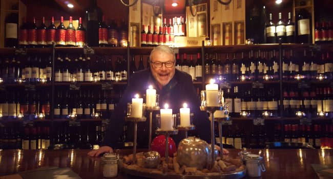 Ein Mann mit Brille, Pius, steht hinter einer Bar, die mit brennenden Kerzen und Dekoartikeln geschmückt ist. Regale voller Weinflaschen bilden den Hintergrund und schaffen eine gemütliche, intime Atmosphäre. Pius lächelt und scheint die Atmosphäre der Umgebung zu genießen.