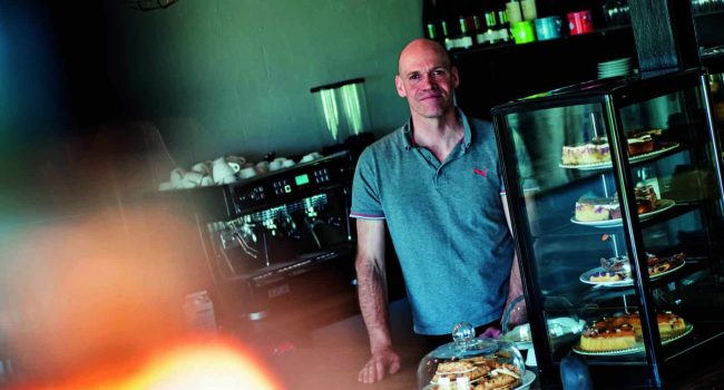 Ein Mann steht in einem gemütlichen Café mit Kaffeerösterei hinter der Theke. Er trägt ein graues Poloshirt. Unter Glasabdeckungen werden auf der Theke verschiedene Gebäcksorten präsentiert. Hinter ihm sind teilweise Tafeln mit Menüs sichtbar, auf denen verschiedene Kaffeespezialitäten aufgelistet sind. Warmes Licht und ein unscharfer Vordergrund schaffen eine einladende Atmosphäre.