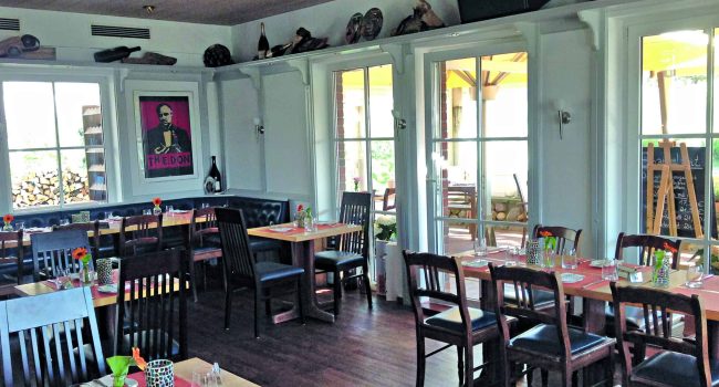 Ein gemütliches Restaurant-Interieur mit ordentlich angeordneten Holztischen und -stühlen. An der Wand neben dem Fenster hängt ein großes Poster von Der Pate. Drei große Fenster sorgen für natürliches Licht. Die Tische sind mit Tischsets, Besteck und kleinen Blumenvasen gedeckt. Dekorative Gegenstände schmücken die Regale über den Fenstern.