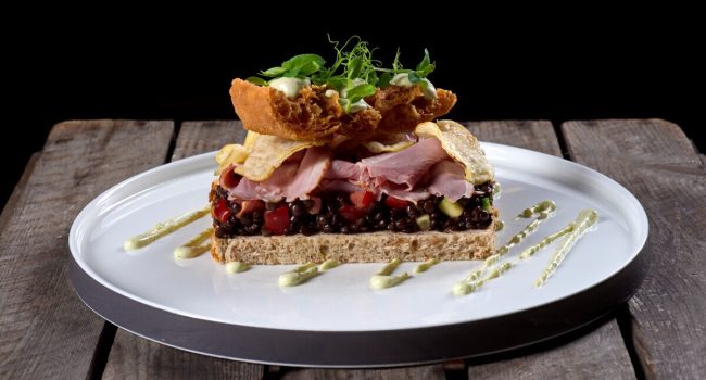 Ein Gourmet-Sandwich liegt auf einem runden weißen Teller mit Holzbrettern im Hintergrund. Das Sandwich, das perfekt zu Brot & Bier passt, besteht aus Schichten von Schinken, Gemüse, Grünzeug und knusprig gebratenen Zwiebeln. Es ist mit einem Schuss Sahnesauce und Microgreens garniert und auf kunstvolle und ansprechende Weise präsentiert.