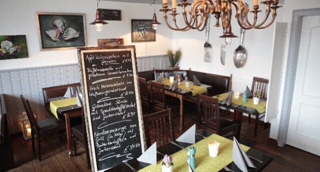 Das gemütliche Restaurant-Interieur bei Janke's besteht aus Holztischen mit gelben Tischdecken und Holzstühlen. In der Mitte steht eine schwarze Tafel mit handgeschriebenen Menüs. Der schwach beleuchtete Raum ist mit Gemälden, einem Kronleuchter und verschiedenen Dekorationsgegenständen an den Wänden geschmückt.