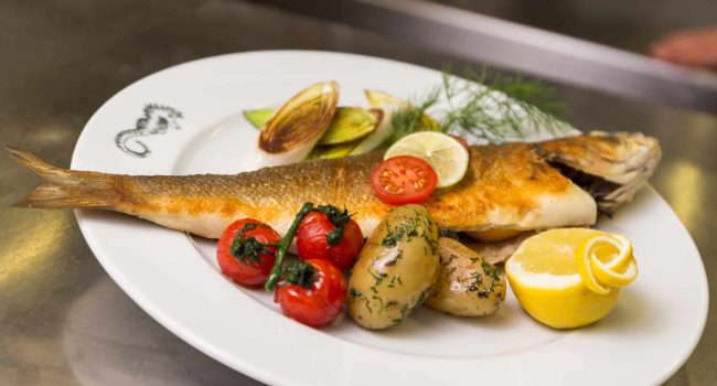 Ein weißer Teller mit einem ganzen gegrillten Fisch, garniert mit einer Limettenscheibe. Zum Fisch gibt es geröstete Kirschtomaten, zwei kleine Kartoffeln, bestreut mit grünen Kräutern, gegrillte Zucchinischeiben und Zitronenscheiben. Ein Zweig Dill ziert den Rand des Tellers und erinnert an die lebhaften Aromen Samoas.