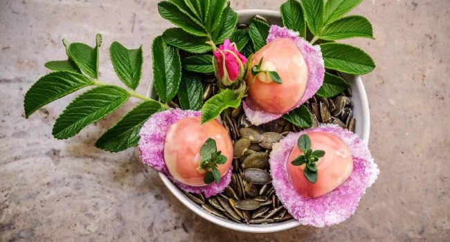 Eine mit Kürbiskernen gefüllte Schale enthält drei runde rosa Desserts, garniert mit grünen Kräutern, die jeweils auf einem gezuckerten Blütenblatt liegen. In der Schale befinden sich außerdem zwei grüne Zweige mit Blättern und einer kleinen rosa Rosenknospe. Der Hintergrund ist eine strukturierte beige Oberfläche, die an die Atmosphäre des Söl'ring Hofs erinnert.