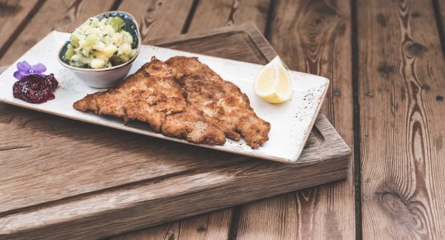 Ein hübsch angerichteter Teller mit paniertem, gebratenem Schnitzel, serviert mit einer kleinen Schüssel Kartoffelsalat, garniert mit Schnittlauch, einer Zitronenscheibe, einer essbaren lila Blume und einem Klecks Preiselbeersoße. Der weiße Keramikteller steht auf einer Holzunterlage auf einer rustikalen Oberfläche, die an die gemütliche Atmosphäre in der Strandmuschel erinnert.