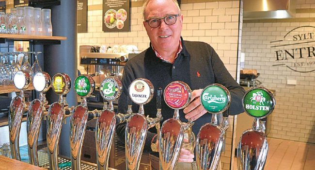 Eine Bar mit einem Barkeeper, der hinter einer Reihe von Zapfhähnen steht. An acht Zapfhähnen sind Marken verschiedener Brauereien zu sehen, darunter Brooklyn, Erdinger, Paulaner, Carlsberg und Holsten. Der Barkeeper trägt eine Brille und einen dunklen Pullover und lächelt in die Kamera. Im Hintergrund sind Regale und eine geflieste Wand zu sehen, die an ein elegantes Entree auf Sylt erinnert.