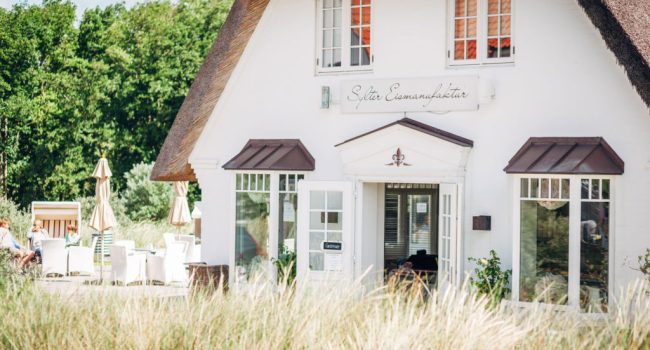 Ein charmantes weißes Häuschen mit Strohdach und großen Fenstern, inmitten von hohem Gras gelegen. Auf der Veranda hängt ein Schild mit der Aufschrift „Sylter Eismanufaktur“. Links sind Sitzplätze im Freien mit beigen Sonnenschirmen zu sehen, die Besucher einladen, köstliches Eis inmitten üppigen Grüns zu genießen.