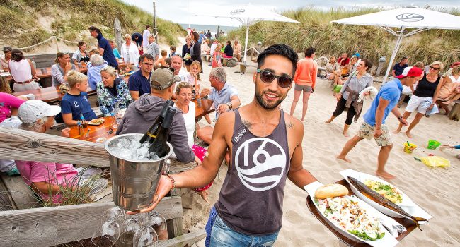 Ein lächelnder Mann mit Sonnenbrille und Tanktop trägt ein Tablett mit Essen und einen Eimer mit Eis, in dem sich eine Weinflasche und Gläser befinden. An der Buhne 16 steht er an einem Sandstrand. Zahlreiche Menschen sitzen an Holztischen oder stehen unter weißen Sonnenschirmen herum. Grasbewachsene Dünen und ein wolkiger Himmel bilden den Hintergrund.