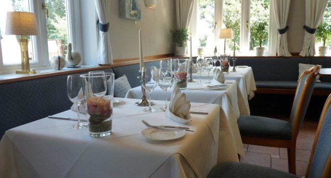 Das gemütliche, sonnendurchflutete Restaurant im Fitschen am Dorfteich bietet elegant gedeckte Tische mit weißer Tischwäsche, gefalteten Servietten und verschiedenen Glaswaren. Auf jedem Tisch steht eine durchsichtige Vase mit dekorativen Steinen und eine Kerze. Große Fenster mit hellen Vorhängen bieten einen Blick auf üppiges Grün. Sanftes Licht trägt zur ruhigen Atmosphäre bei.