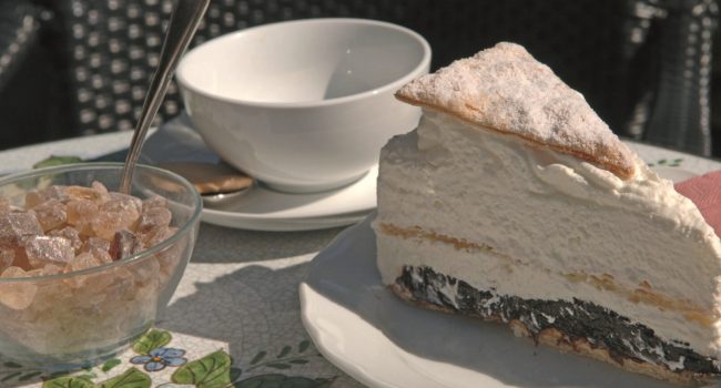 Ein Stück Schichtcremetorte mit einer Prise Puderzucker liegt auf einem Teller neben einer leeren weißen Schüssel und einer Untertasse mit einem Löffel. Daneben steht eine kleine Schüssel mit braunem Kandiszucker und einem Metalllöffel. Das Gedeck, das an eine Kleine Teestube erinnert, steht auf einem mit Mosaikfliesen und Blumenmustern geschmückten Tisch.