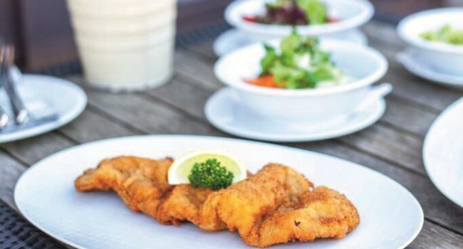 Ein weißer Teller mit drei Stücken goldbraun gebratenem Fisch, garniert mit einer Zitronenscheibe und einem Zweig Petersilie, steht auf einem Holztisch in der charmanten Kleinen Küchenkate. Im Hintergrund stehen drei unscharfe weiße Schüsseln mit grünen Salaten mit verschiedenen Gemüsesorten.