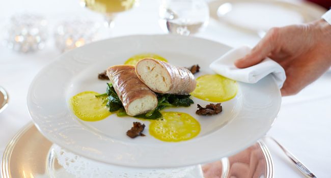 Eine Hand mit einer weißen Serviette stellt im Miramar einen eleganten Teller mit Gourmet-Essen auf eine makellos weiße Tischdecke. Das Gericht besteht aus zwei gerollten Fleischstücken auf einem Bett aus sautiertem Gemüse, garniert mit gelber Soße und kleinen, dunklen essbaren Akzenten. Im Hintergrund stehen Gläser mit Weißwein und Wasser.