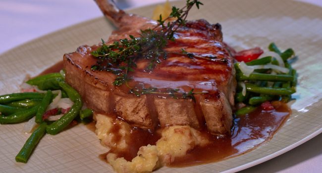 Ein gegrilltes Schweinekotelett, garniert mit frischem Thymian, wird auf einem Bett aus cremigem Kartoffelpüree mit sautierten grünen Bohnen und Zwiebeln serviert. Beträufelt mit einer reichhaltigen braunen Soße wird das Gericht auf einem hellen Teller präsentiert und wird jeden Gast beeindrucken, sogar Klaus selbst.