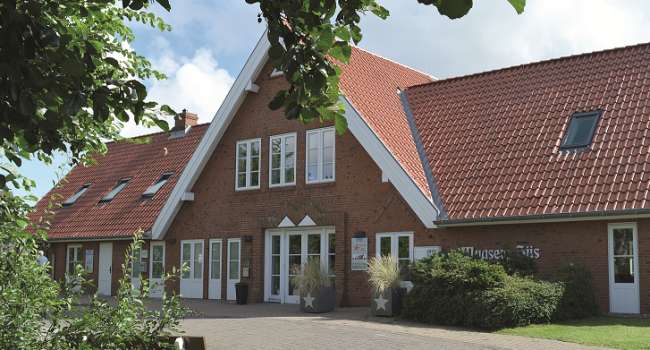 Ein großes Gebäude aus rotem Backstein mit steilem Dach aus roten Ziegeln und weiß gerahmten Fenstern unter blauem Himmel. Der Eingang ist von großen Topfpflanzen flankiert und von einer gepflegten Landschaft mit Bäumen und einer Hecke umgeben. Das Schild „Haus am Meer – Haus am Sils“ ist teilweise sichtbar und vermittelt das Gefühl, „wie zuhause“ zu sein.